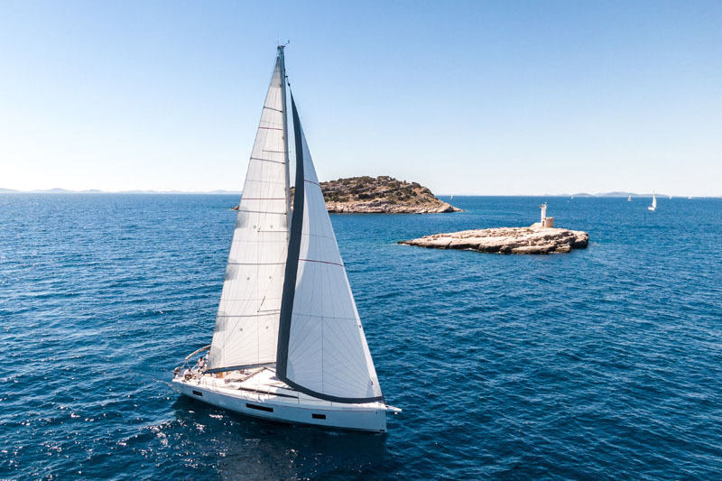 Photo has sailing yach with open sails in front of small island with lighthouse. It is nice sunny day without clouds in a sky.